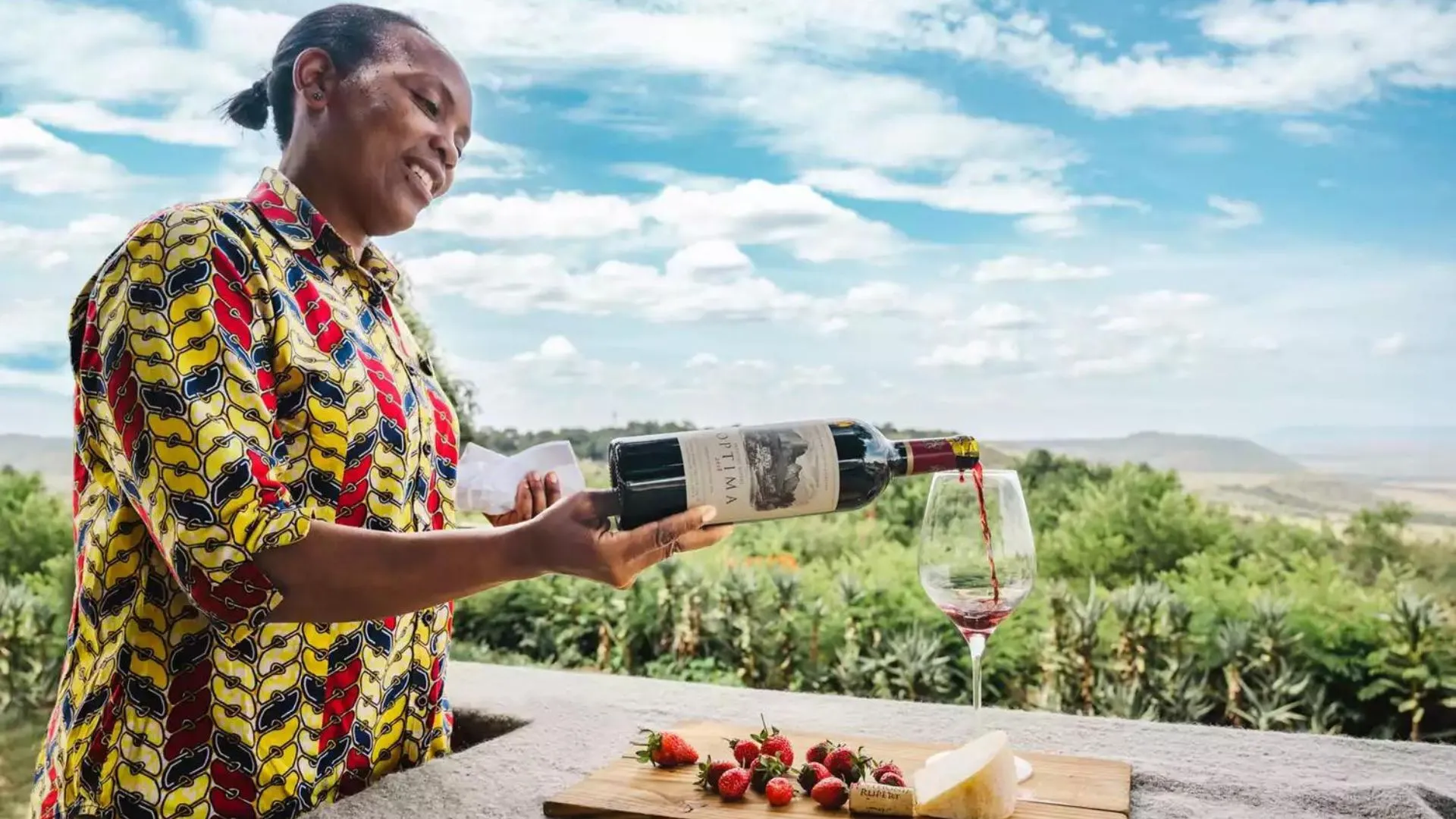 A woman pours red wine into a glass on a terrace overlooking green countryside.